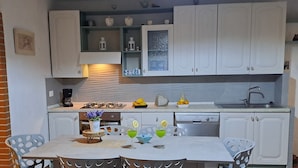 Une cuisine compacte avec des meubles blancs, une cuisinière à gaz et un lave-vaisselle. On y trouve une table à manger avec quatre chaises et une petite plante en pot.
