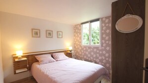 A bedroom with a bed, two bedside tables with lamps, a window with floral wallpaper, and a door with a round mirror.
