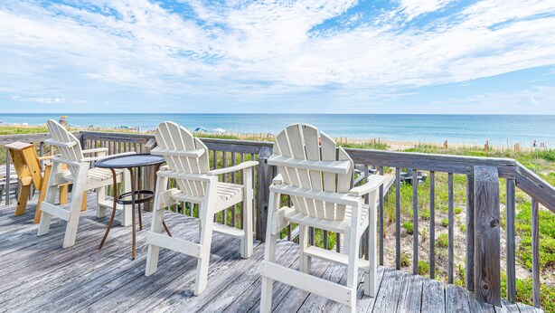 A wooden deck with white Adirondack chairs and a small round table, overlooking a beach with a wooden railing.