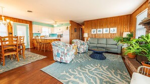 A cozy living room with a sofa, chairs, and a coffee table. There is a dining area with a table and chairs. The room has wooden walls and a ceiling fan.