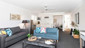 Ein modernes Wohnzimmer mit einer grauen Couch, einer blauen Couch und einem Couchtisch. Im Hintergrund ist eine Küche mit weißen Schränken und einer Mikrowelle zu sehen.