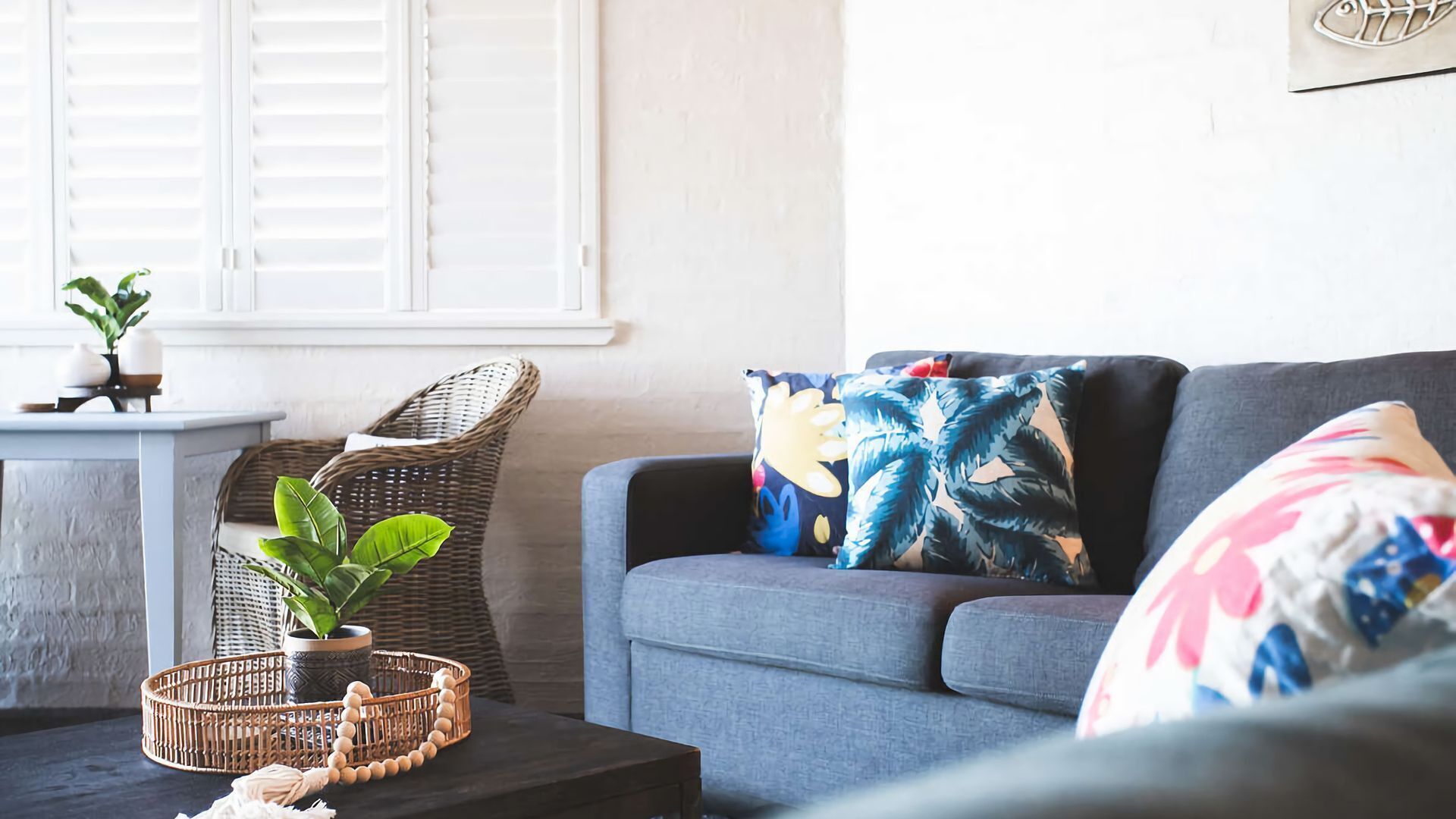 A living room with a grey sofa, a wicker chair, a small table with a plant, and a decorative pillow with a tropical leaf pattern.