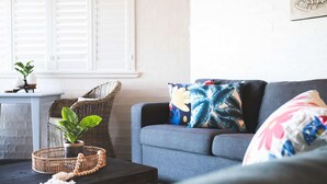 Ein Wohnzimmer mit grauem Sofa, einem Korbstuhl, einem kleinen Tisch mit Pflanze und einem dekorativen Kissen mit tropischem Blattmuster.