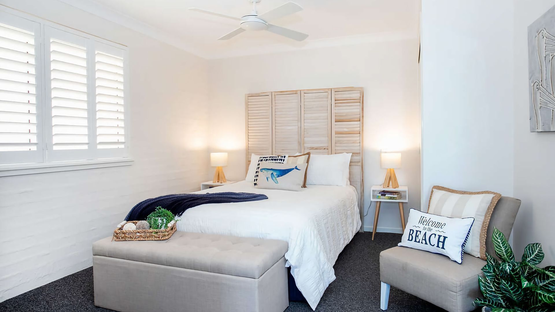 A bedroom with a bed, bedside tables, a ceiling fan, a window with blinds, and a wall-mounted artwork.