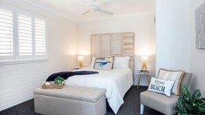 Ein Schlafzimmer mit Bett, Nachttischen, Deckenventilator, Fenster mit Jalousien, Nachttischlampe, Zierkissen, einer kleinen Pflanze und einem Sessel mit Kissen.