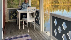 Une véranda couverte avec une table et des chaises en bois, un tapis à motifs et une vue sur un lac entouré d’arbres.