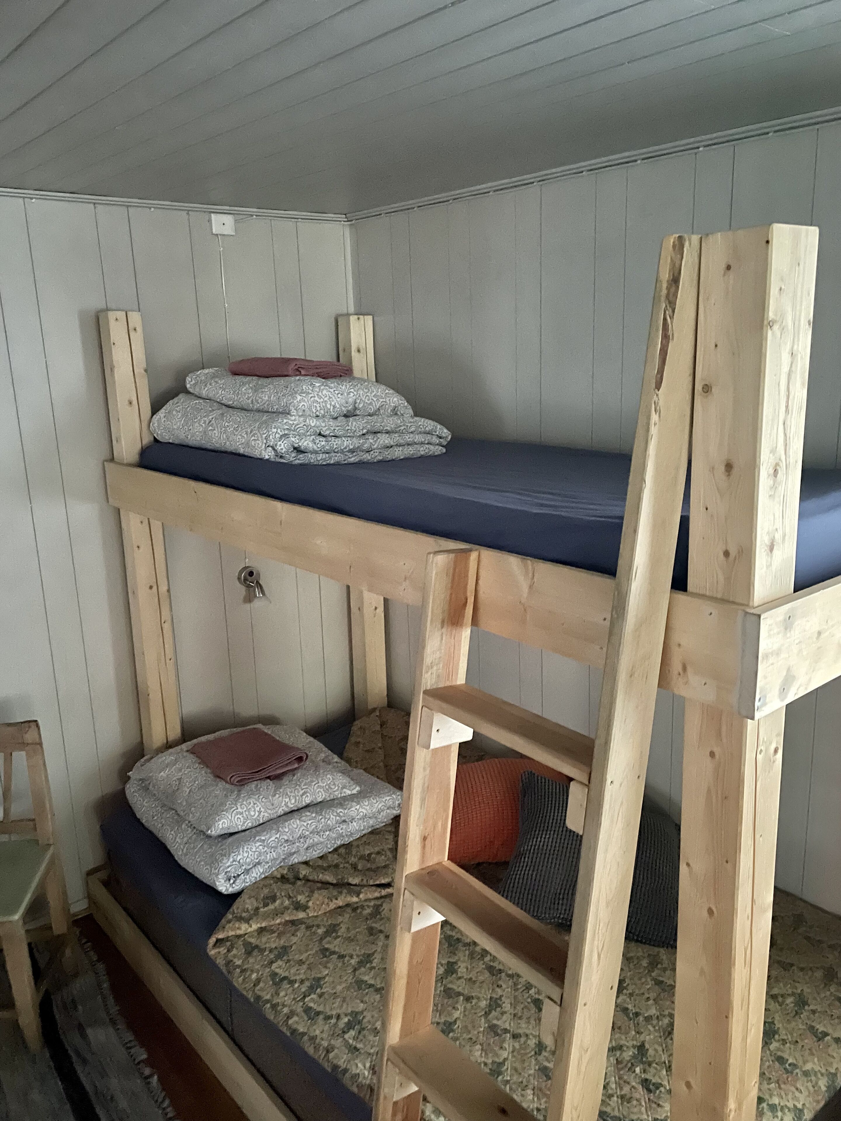 En våningssäng med en trästege, ett litet bord och en stol i ett rum med träväggar och en mönstrad matta.