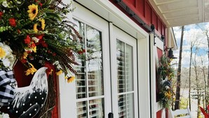 Une porte blanche avec des panneaux de verre, flanquée d’une décoration représentant un coq et d’un grand arrangement floral.