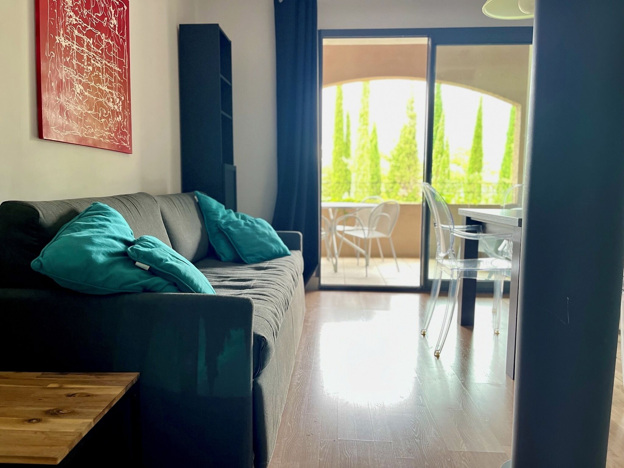 A modern living room with a grey sofa, teal cushions, a wooden coffee table, and a large window offering a view of trees and a patio.