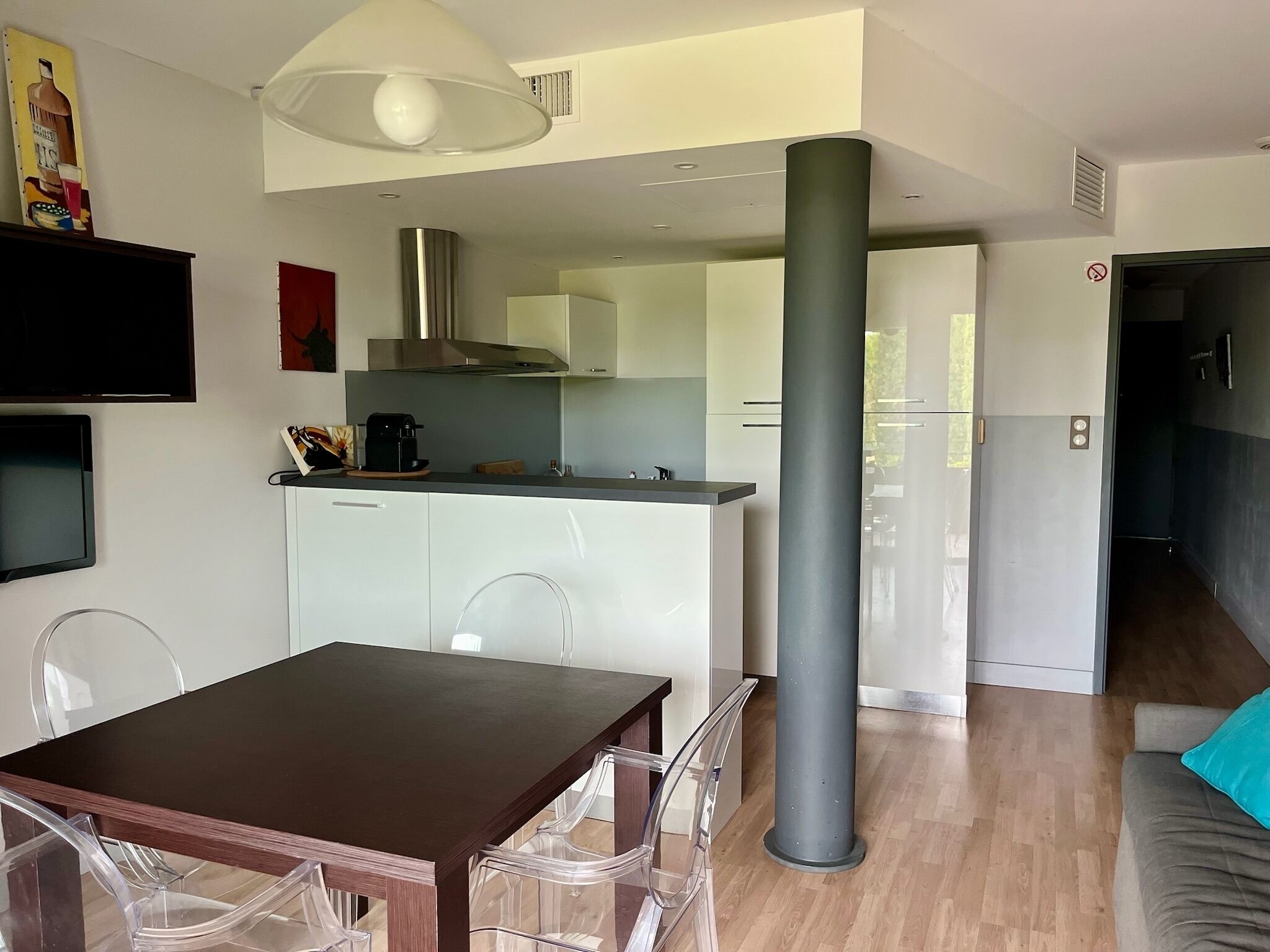 A modern kitchen with white cabinets, a stainless steel range hood, and a coffee maker. There is a dining table with clear chairs and a grey pillar in the center.