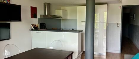 A modern kitchen with white cabinets, a stainless steel range hood, and a coffee maker. There is a dining table with clear chairs and a grey pillar in the center.