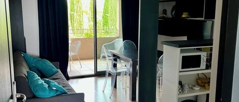 A modern living room with a grey sofa, a wooden bench, and a dining area with a table and chairs. There is a balcony with a view of greenery.