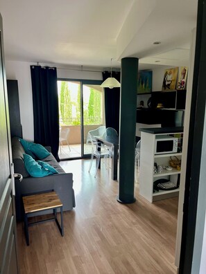 A modern living room with a grey sofa, a wooden bench, and a dining area with a table and chairs. There is a balcony with a view of greenery.