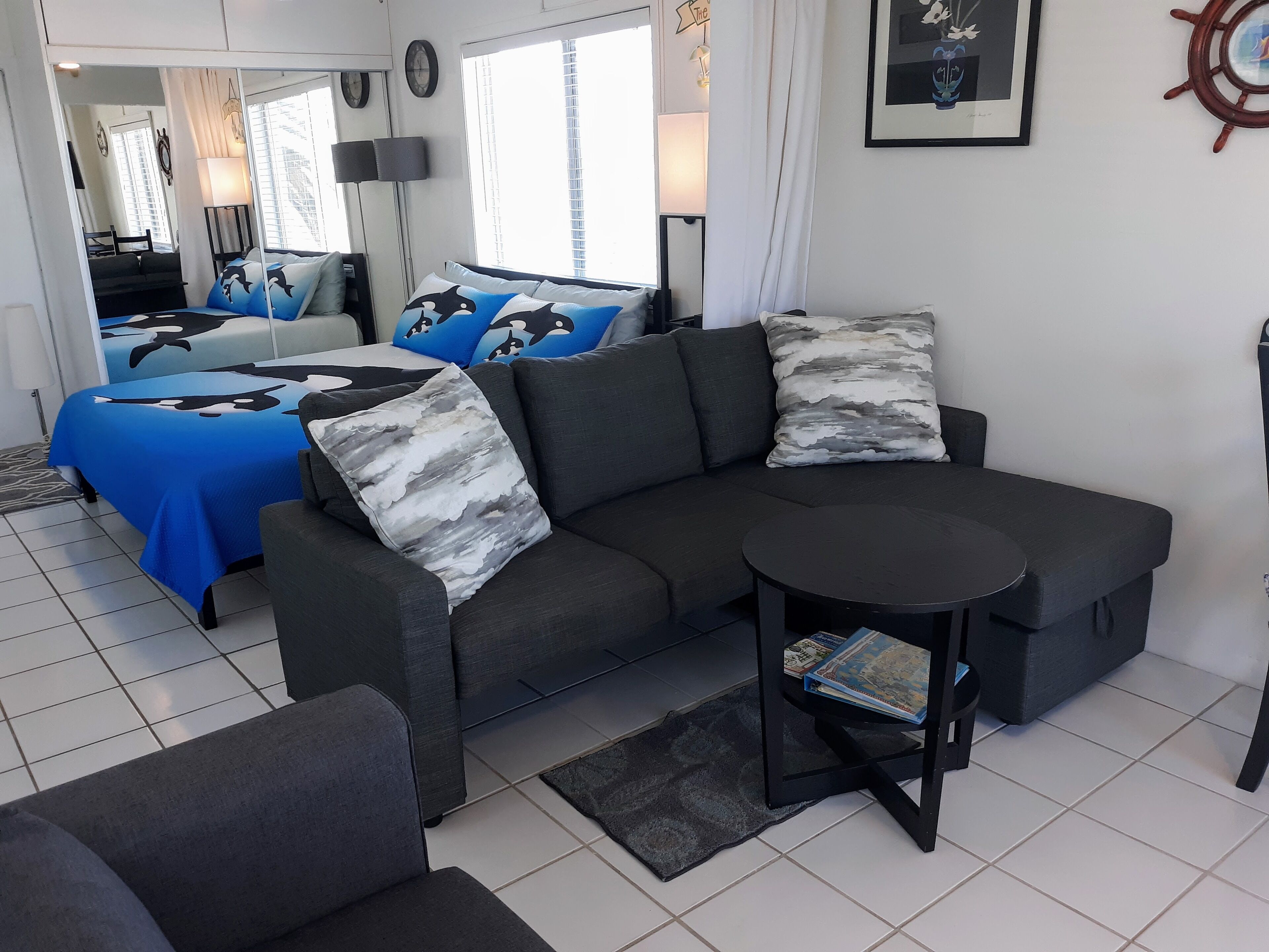 Un salon moderne avec un canapé gris, une table basse ronde et un lit avec une literie bleue et blanche.