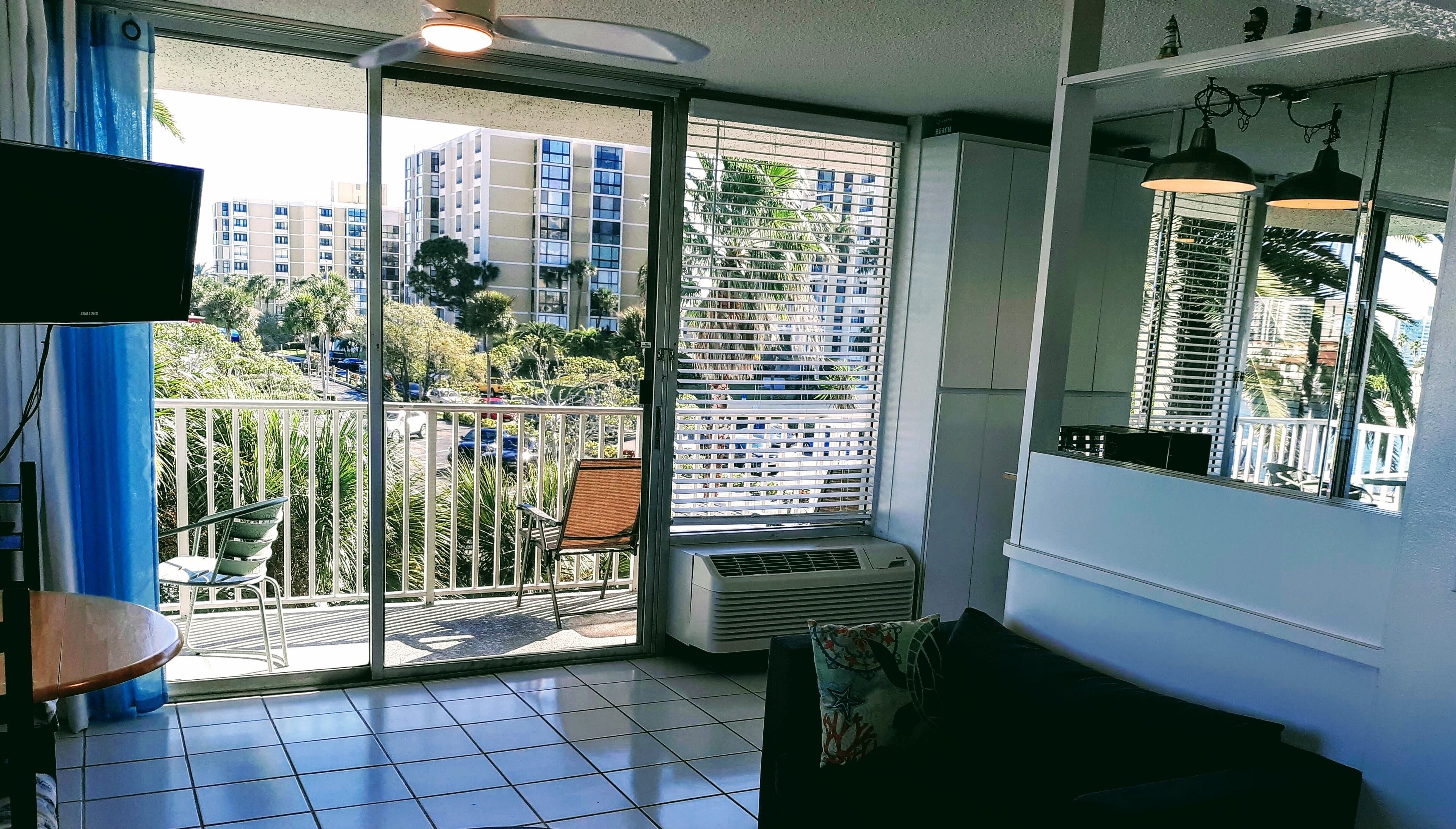 A balcony with a view of tall buildings and parked cars, a sliding glass door, and a ceiling fan.
