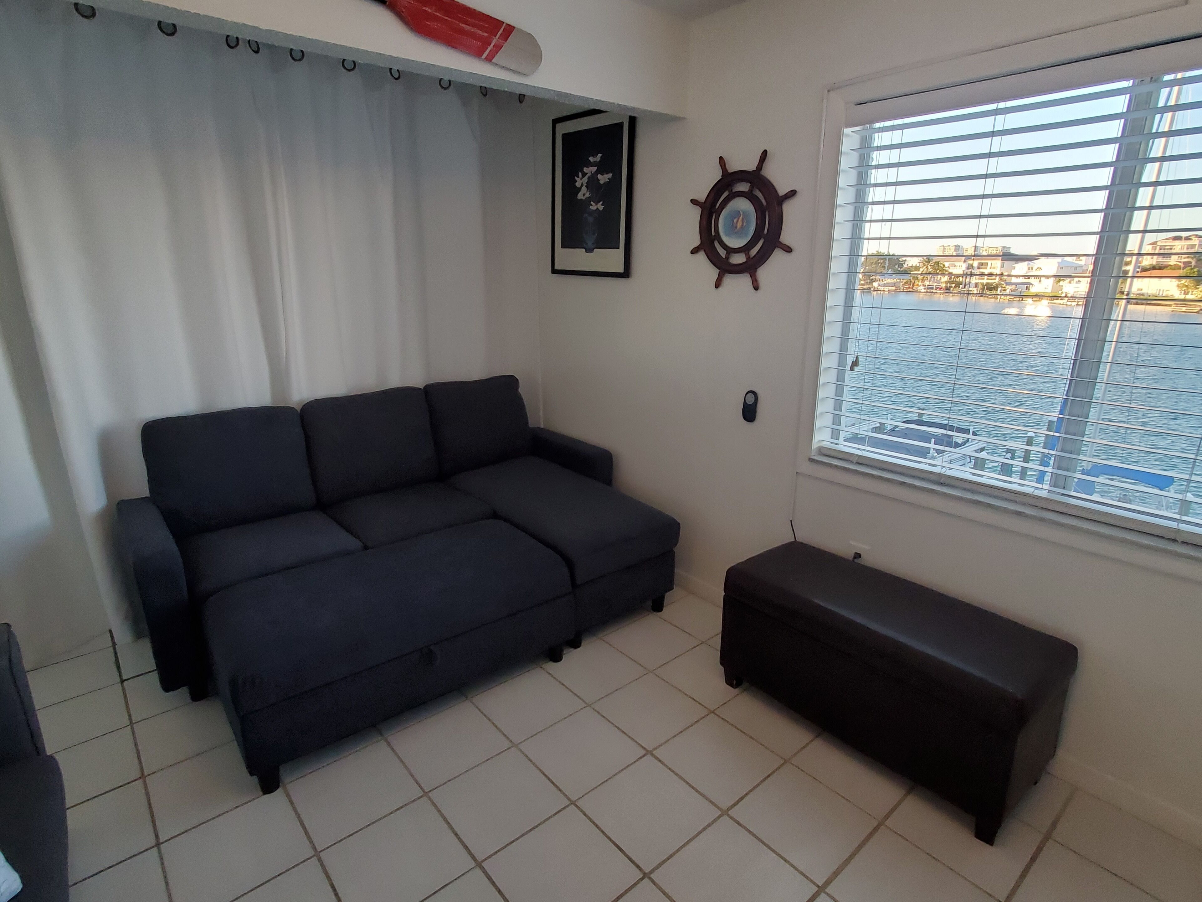 Una sala de estar con un sofá oscuro, un banco, un ventanal con persianas y un timón de barco montado en la pared, además de un cuadro enmarcado.