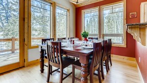 Uma sala de jantar com mesa de madeira posta para quatro pessoas, janelas amplas e vista para árvores.