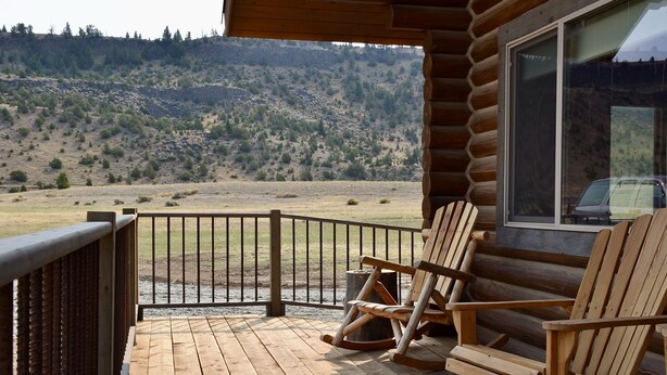 A wooden deck with two Adirondack chairs, a view of a hilly landscape, and a cabin with a large window. - River Haven Cabin-South-Private River Access! (Emigrant)