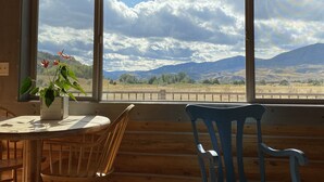 A wooden cabin interior with a dining table, chairs, and a window offering a view of mountains and a cloudy sky. - River Haven Cabin-South-Private River Access! (Emigrant)