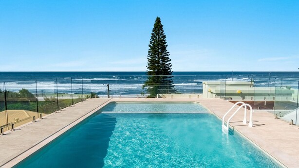 Une piscine rectangulaire à l’eau d’un bleu limpide, entourée d’une terrasse carrelée. Un grand conifère se dresse au centre, et l’océan est visible au loin.