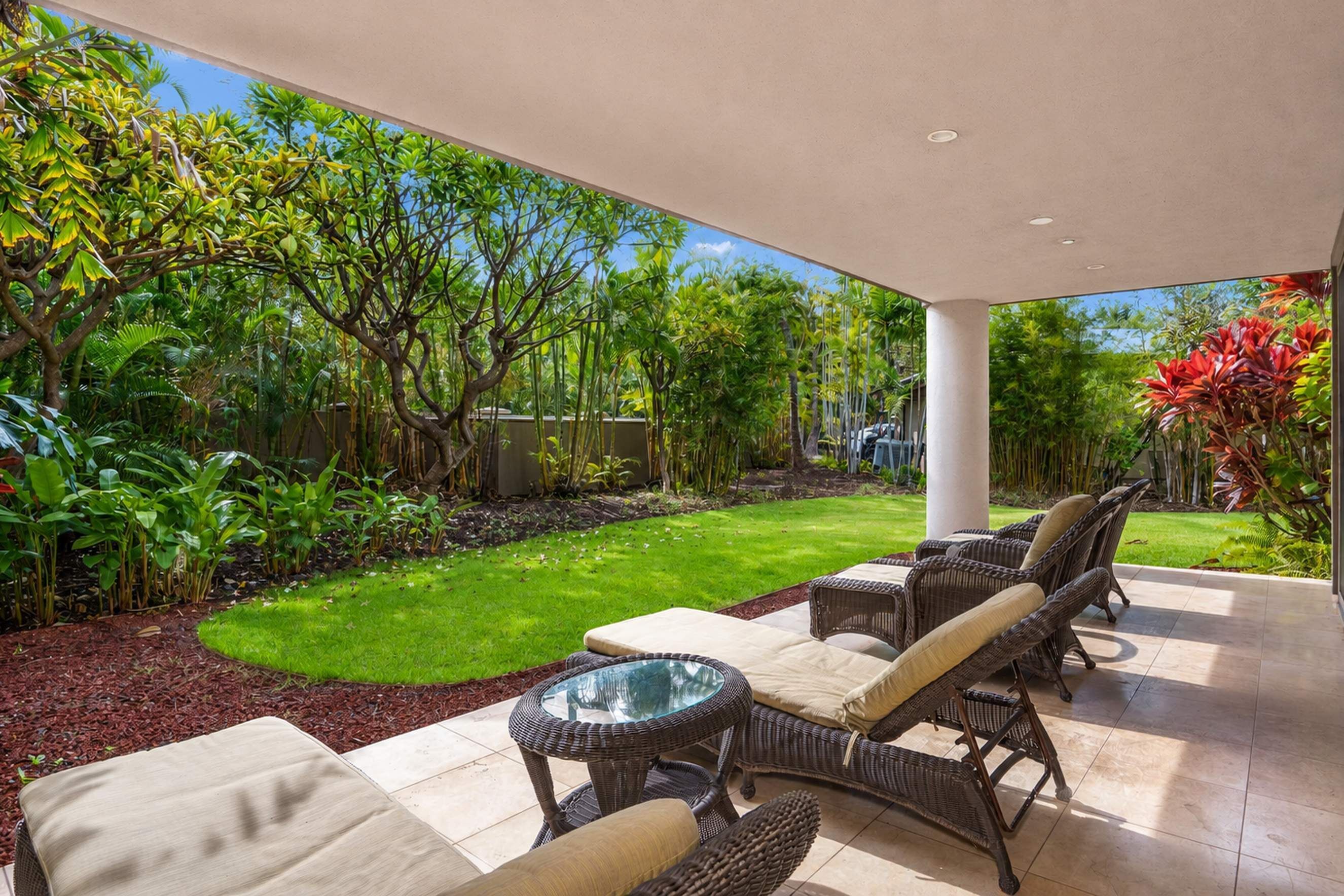 A covered patio with wicker furniture and a glass table, surrounded by a lush garden with various plants and a well-maintained lawn.
