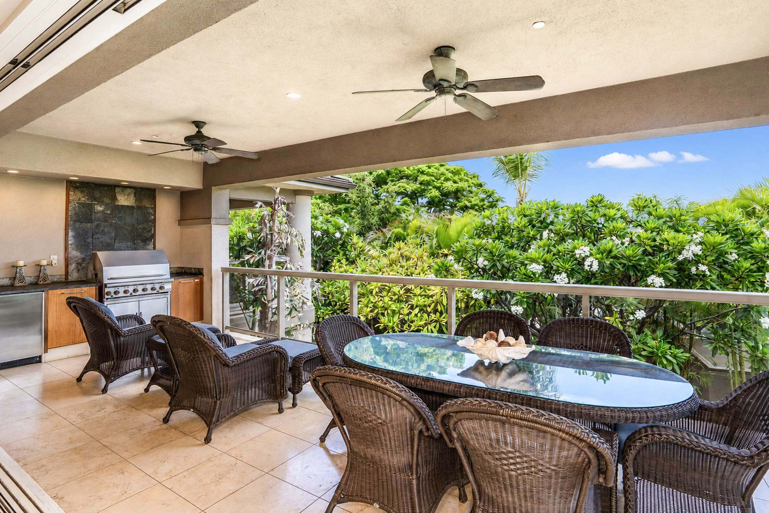 A covered outdoor dining area with wicker furniture, a glass table, and a view of lush greenery.