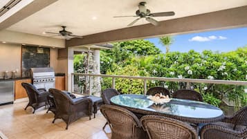 A covered outdoor dining area with wicker furniture, a glass table, and a view of lush greenery.