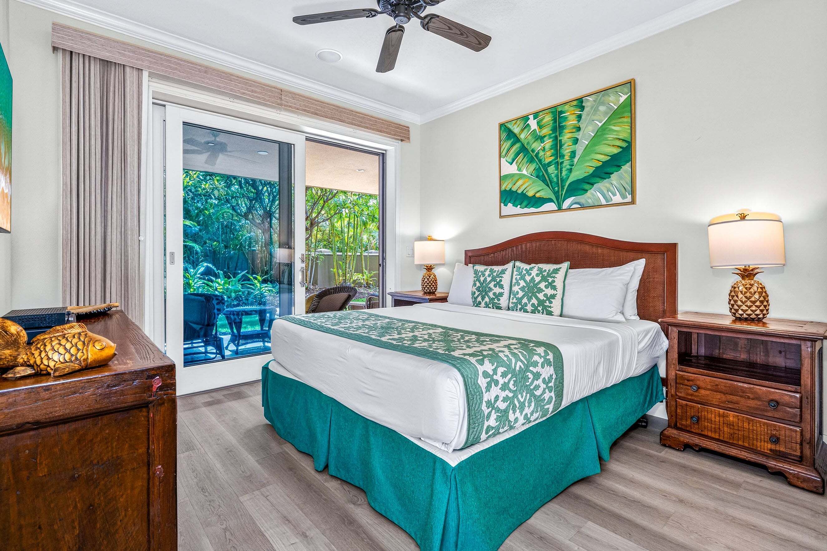 Une chambre d’hôtel avec un lit, un ventilateur de plafond, une fenêtre donnant sur un paysage verdoyant et une œuvre d’art accrochée au mur.