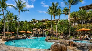 A pool area with a clear blue pool, palm trees, and a rocky waterfall feature. - Luxurious Ho'olei 12-2, Lush View, Close to Pool, Sleeps 8 in beds, Central AC! (Kihei)