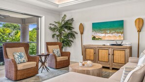 A spacious living area with wicker furniture, a large painting of a beach, and a view of lush greenery. - Luxurious Ho'olei 12-2, Lush View, Close to Pool, Sleeps 8 in beds, Central AC! (Kihei)