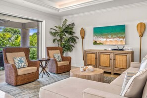 A spacious living area with wicker furniture, a large painting of a beach, and a view of lush greenery. - Luxurious Ho'olei 12-2, Lush View, Close to Pool, Sleeps 8 in beds, Central AC! (Kihei)