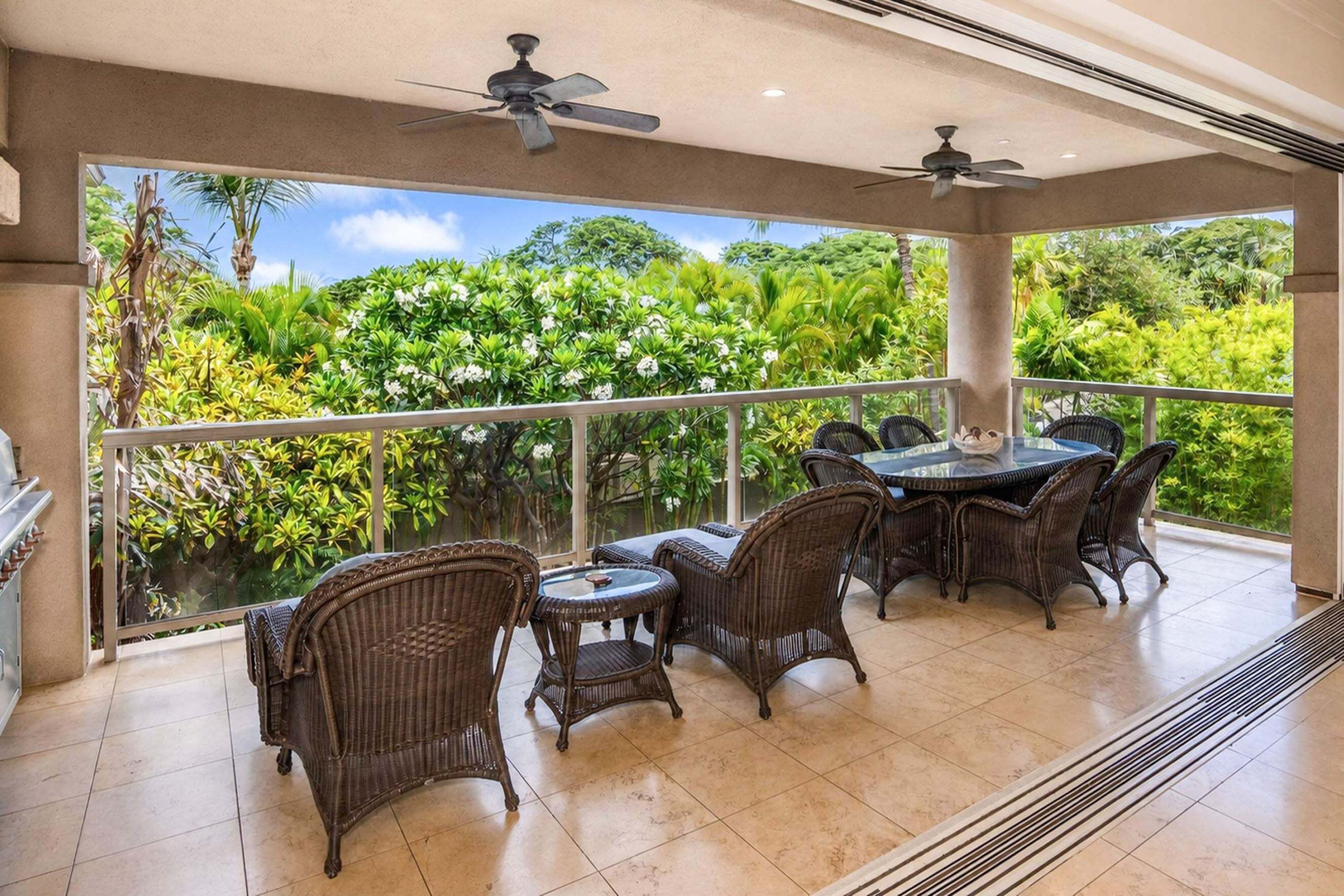 A covered outdoor dining area with wicker furniture and ceiling fans, overlooking a lush garden.