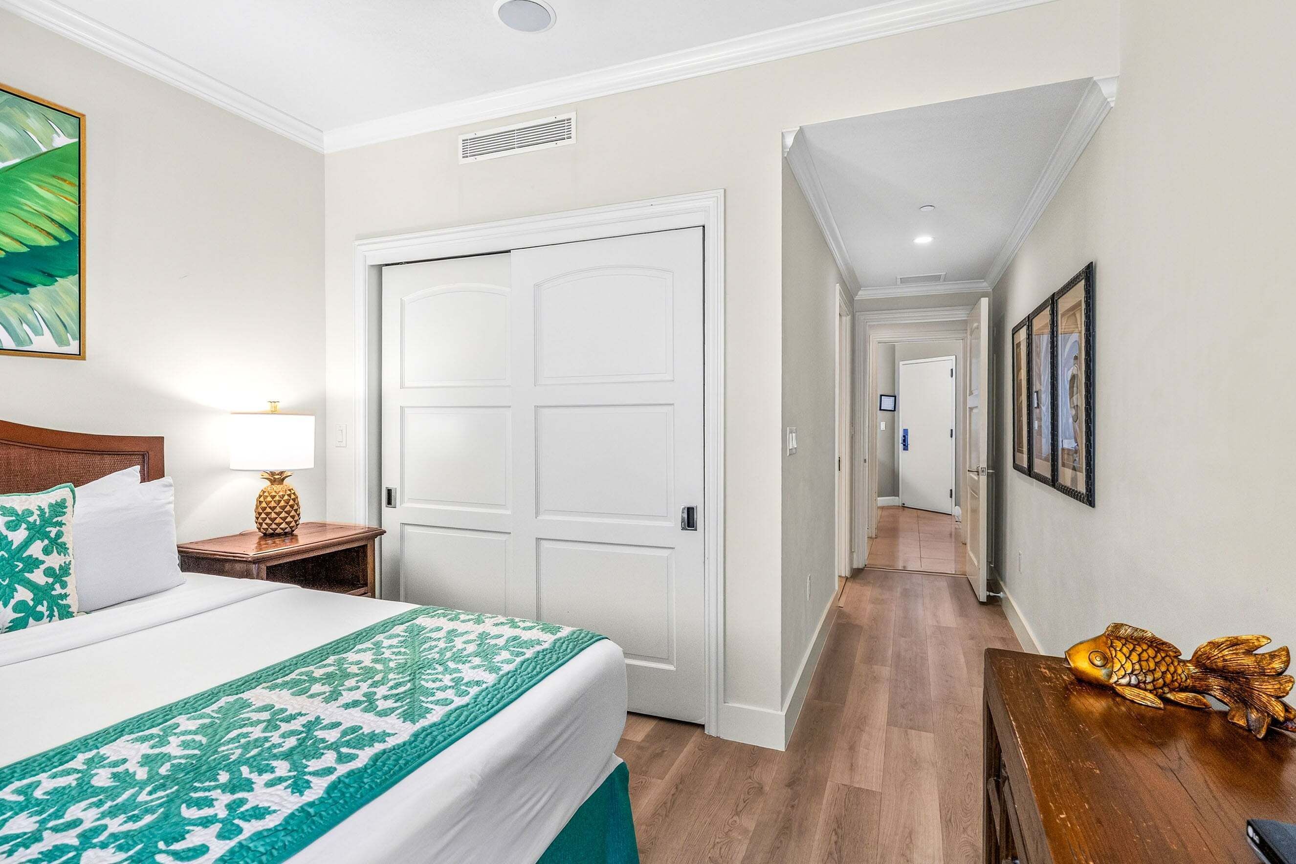 A hotel room with a bed, a nightstand, a lamp, a door, and a hallway leading to another room.