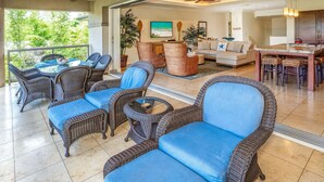 A spacious living area with wicker furniture, a dining table, and a kitchen area. - Luxurious Ho'olei 12-2, Lush View, Close to Pool, Sleeps 8 in beds, Central AC! (Kihei)