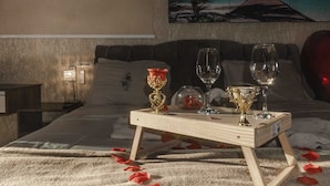 Un dormitorio con una cama, una mesita de noche con una bandeja que sostiene copas de vino y una vela, y un cuadro con una escena de playa en la pared.