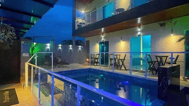 Zona de piscina con barandilla de vidrio, mobiliario exterior y un edificio con varias ventanas y puertas.