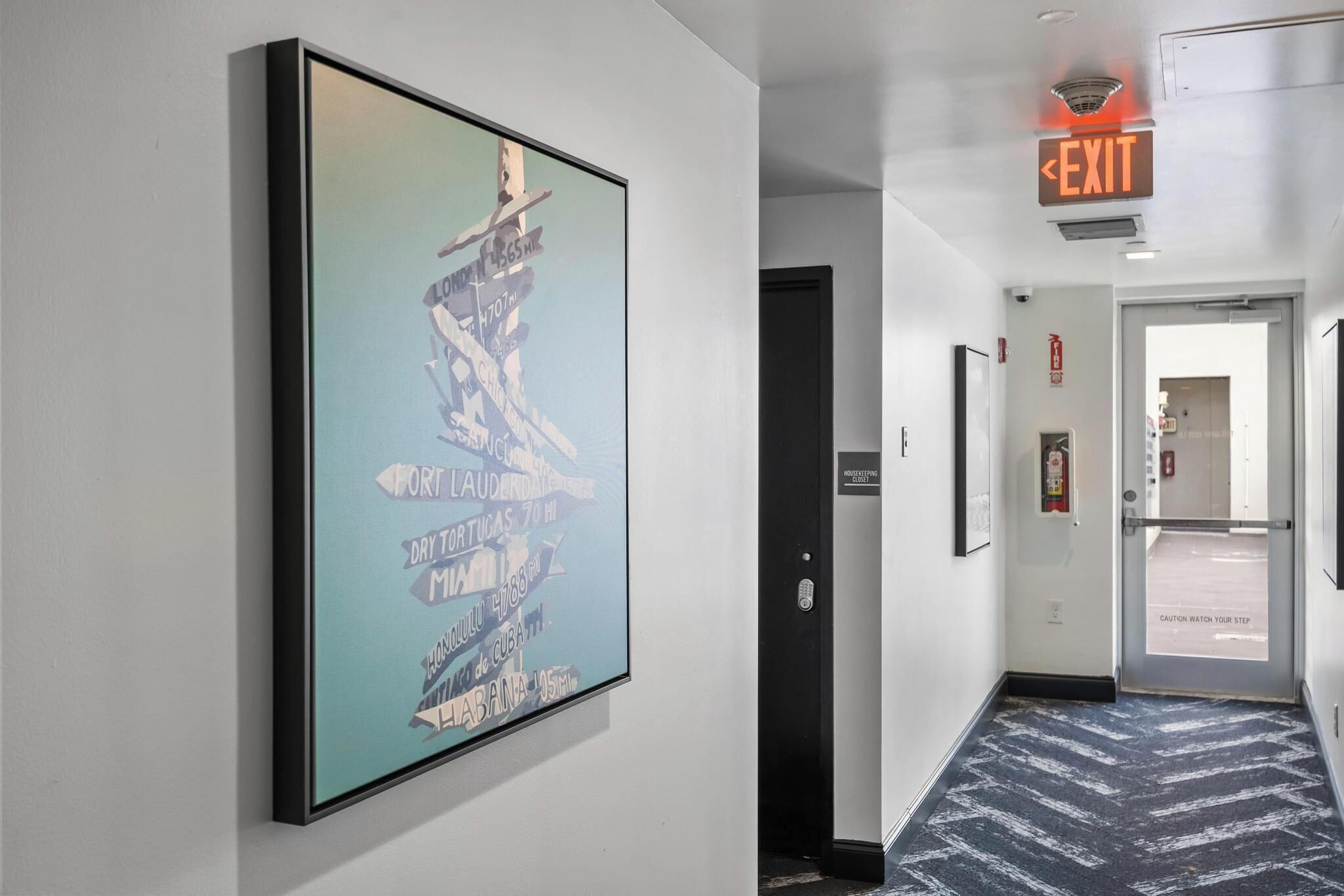 A hallway with a framed map on the wall, an exit sign, and a caution sign on the floor.