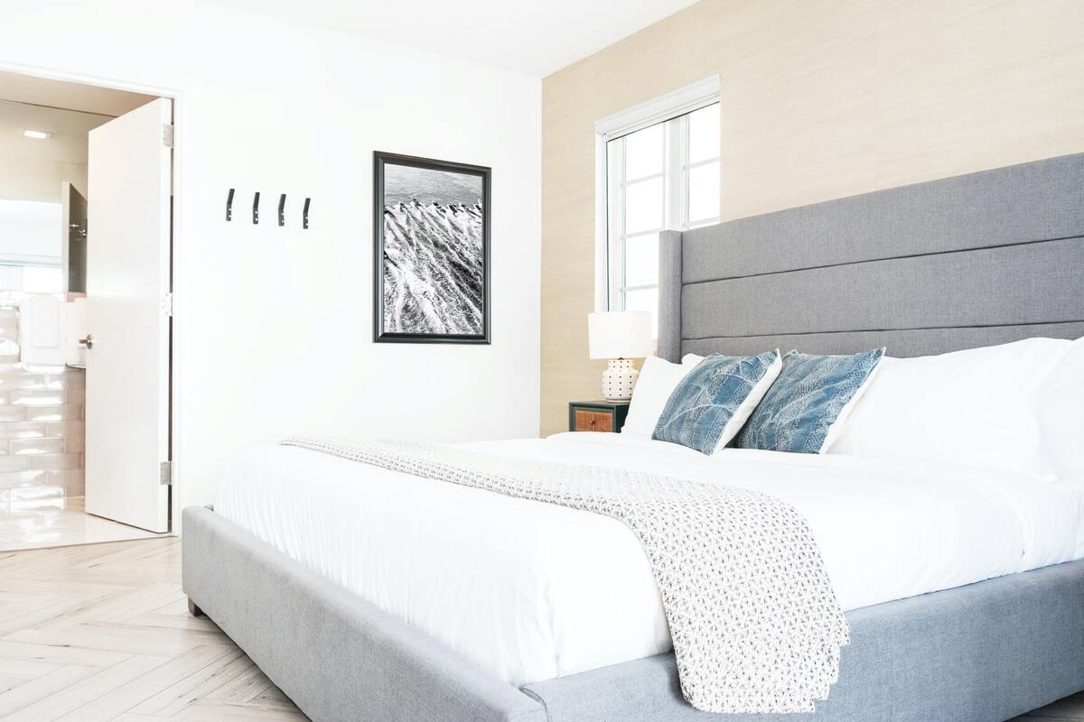 Une chambre à coucher moderne avec un grand lit, une tête de lit grise, une table de chevet avec une lampe et un tableau encadré au mur.