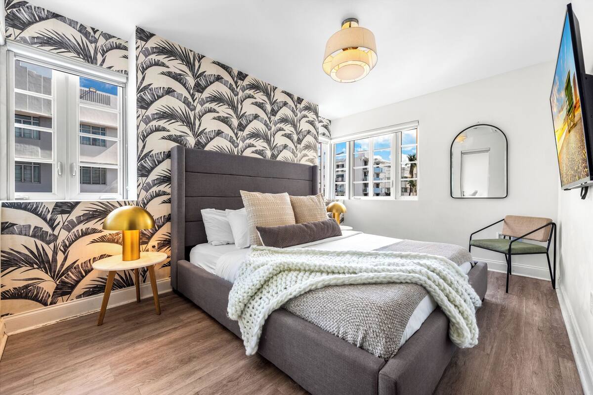 Une chambre moderne avec un grand lit, une vue sur les immeubles par la fenêtre, un miroir rond et une petite table avec une lampe.