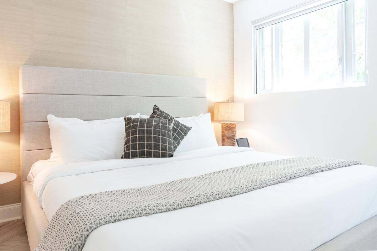 Un lit bien fait, avec des oreillers blancs et à motifs, une tête de lit grise et une lampe de chevet.