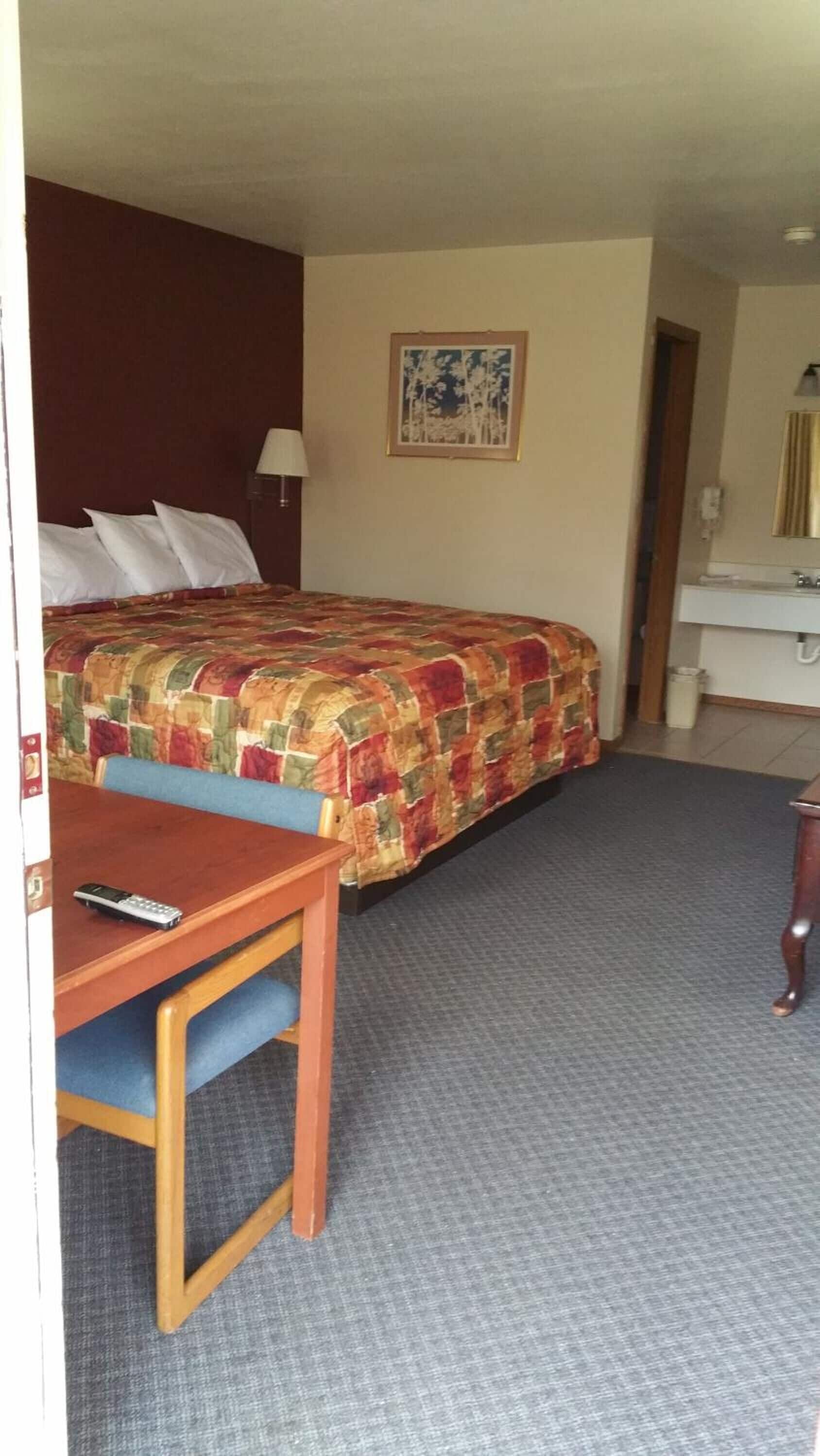 Ett hotellrum med en säng, ett skrivbord med en stol, en lampa, en inramad tavla på väggen och ett badrum med handfat och spegel.