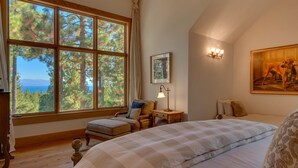 Un dormitorio con un ventanal que ofrece vistas a árboles y un cuerpo de agua. Hay una cama con colcha blanca a cuadros, una silla con cojines, una mesita de noche con lámpara, y un cuadro en la pared.