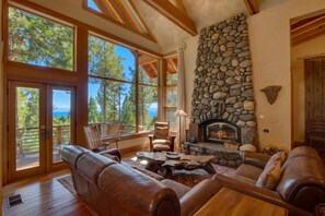 Relax in a spacious living area with a stone fireplace and a large window offering a view of the surrounding forest and water.