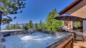 Un jacuzzi con cubierta de madera y mobiliario exterior, con vistas a un lago y montañas al fondo.