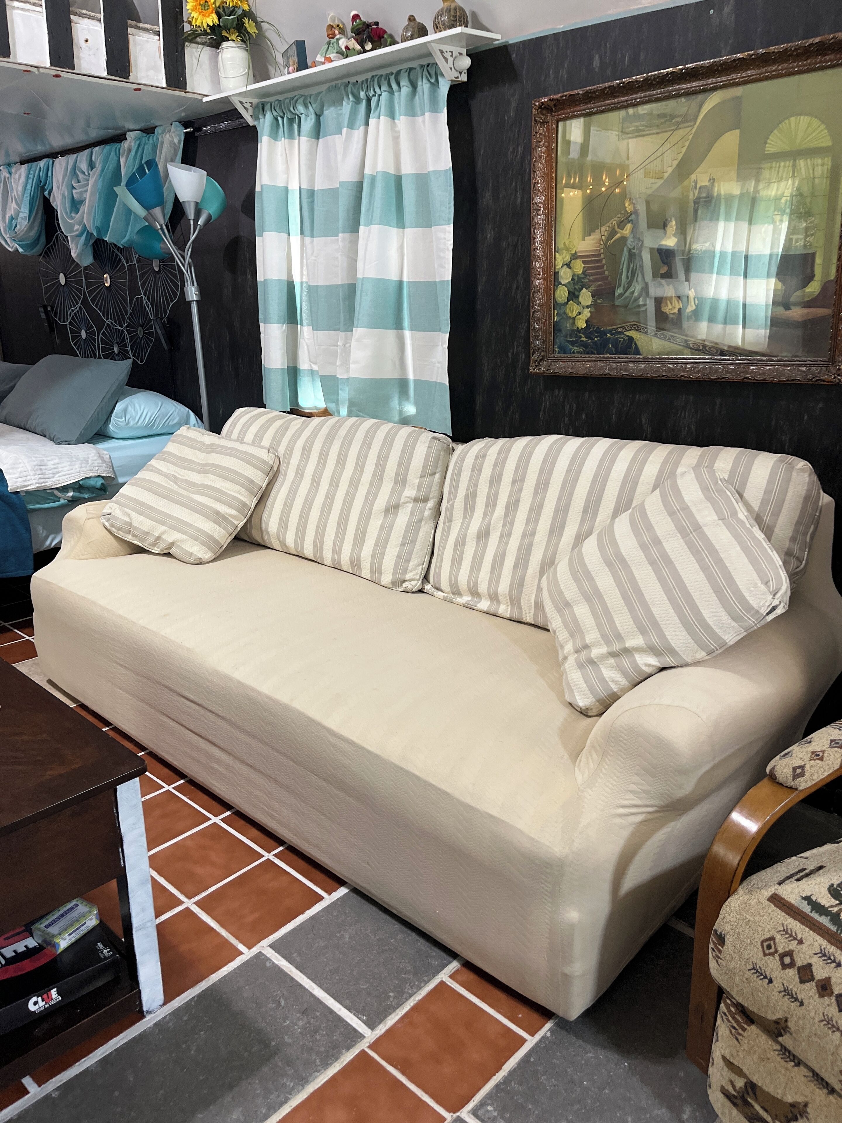 A beige sofa with striped pillows, a tiled floor, a dark wall, a ceiling fan, and a shelf with decorative items.