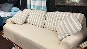 A beige sofa with striped pillows, a tiled floor, a dark wall, a ceiling fan, and a shelf with decorative items.