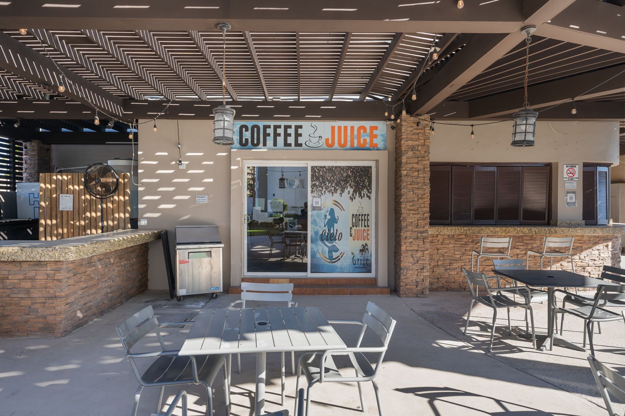 Un área de estar al aire libre con mesas y sillas, un cartel de café y zumos, y una pared de ladrillo.