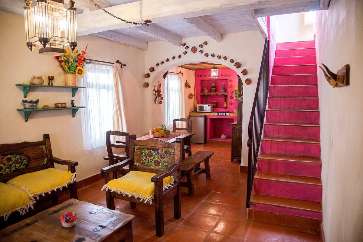 A cozy living room with wooden furniture, a dining area, and a staircase leading to a pink kitchen.