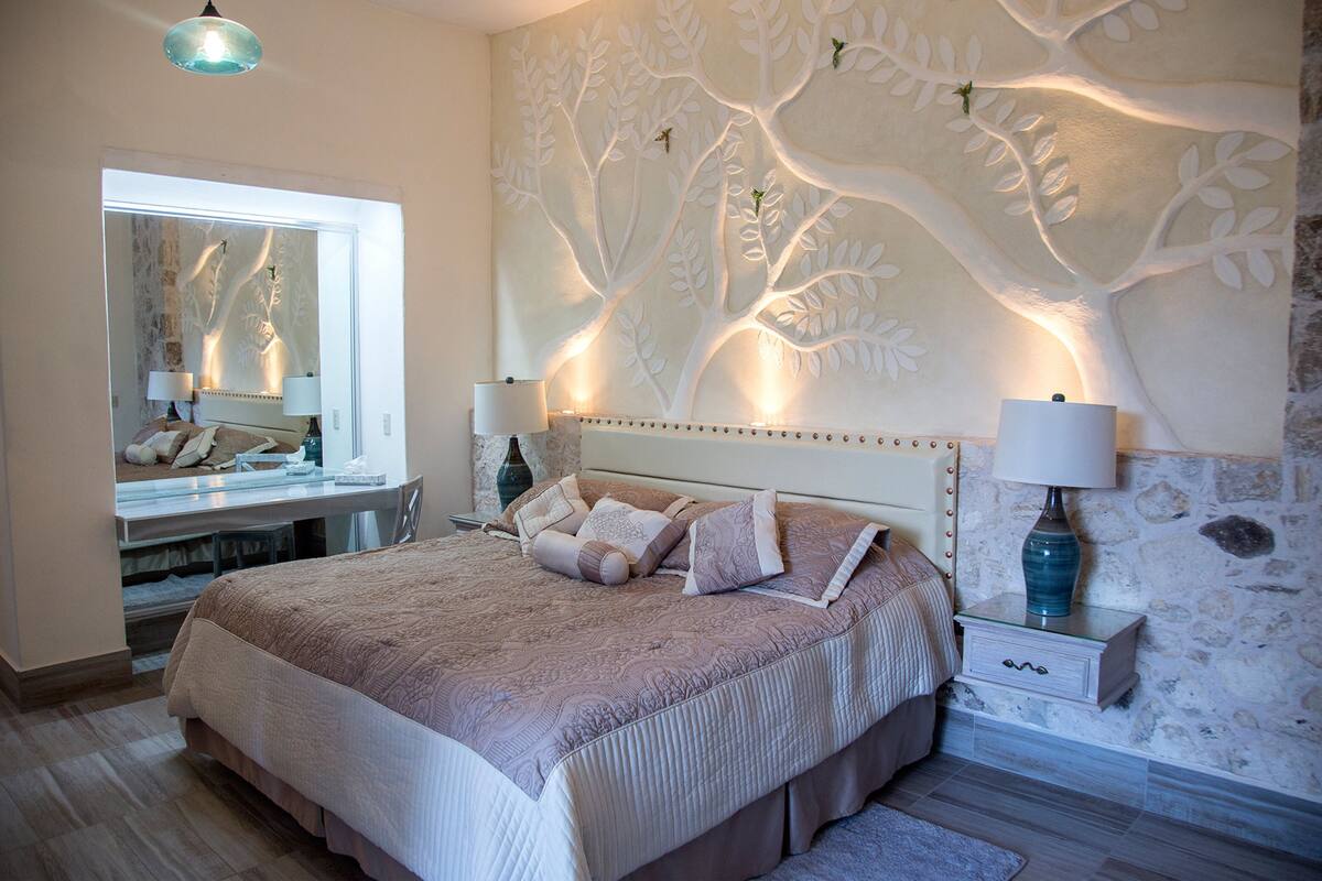 A bedroom with a large bed, a nightstand with a lamp, a dresser, and a mirror.