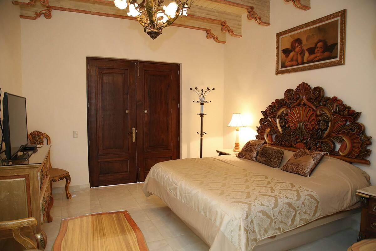 A bedroom with a large bed, a wooden wardrobe, a desk with a chair, a lamp, and a painting on the wall.